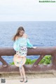 A woman in a blue top and floral skirt leaning on a railing by the ocean.