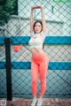 A woman in a white top and orange leggings posing in front of a fence.